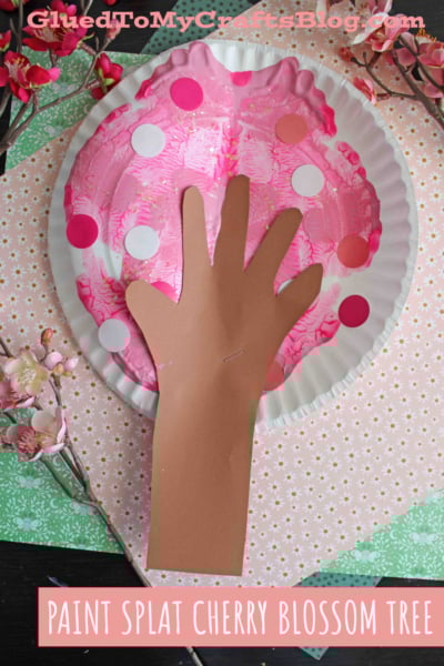 A craft project featuring a paper plate painted pink with white and pink dots to resemble cherry blossoms, and a brown paper hand cutout attached to represent a tree trunk and branches. Text reads PAINT SPLAT CHERRY BLOSSOM TREE.