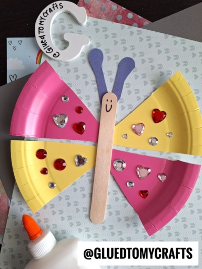 A butterfly craft made from pink and yellow painted paper plate pieces, decorated with heart-shaped gems, a craft stick body with a smiley face, purple antennae, and a bottle of glue nearby. @GLUEDTOMYCRAFTS is labeled.