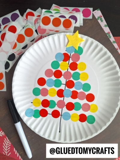 A paper plate decorated with colorful dot stickers arranged in a Christmas tree shape, topped with a yellow star. Beside the plate are a black marker and sheets of coding label crafts. The username @GLUEDTOMYCRAFTS is at the bottom.