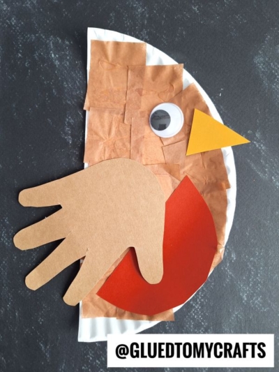 A children’s craft of a bird made from a paper plate, brown paper, a red paper semi-circle, a yellow paper beak, a googly eye, and a handprint cutout for the wing. @GLUEDTOMYCRAFTS is at the bottom.