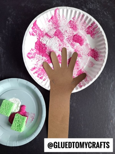A paper plate with pink and white sponge paint, a brown paper cutout of an arm and hand glued on, and a smaller plate with two sponge pieces—perfect for cherry blossom tree crafts. @GLUEDTOMYCRAFTS text is at the bottom.