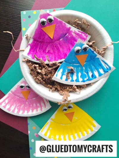Four colorful paper plate bird crafts, each with big googly eyes and an orange beak, are displayed on a multicolored background. Three brighten the scene outside while one sits playfully in a bowl with brown paper shreds.