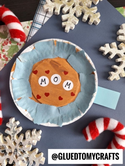 A handmade paper craft resembling a mug of hot chocolate with a coding label reading MOM, surrounded by felt hearts, blue tissue, fake snowflakes, and striped candy canes. The handle is made from blue paper. @GLUETOMYCRAFTS is written at the bottom.