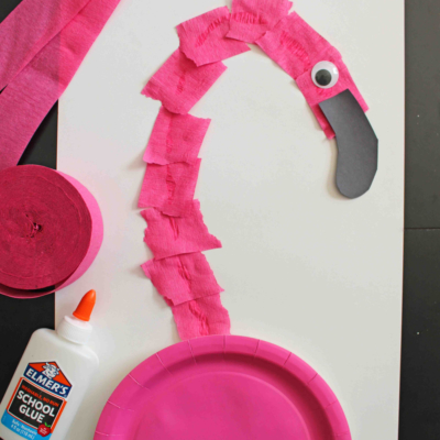 A fun craft project featuring a torn crepe paper flamingo, with a pink paper plate body, black paper beak, googly eye, and white glue, all arranged on a white background.