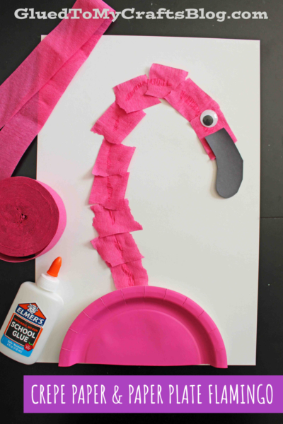 A fun craft project featuring a torn crepe paper flamingo, with a pink paper plate body, black paper beak, googly eye, and white glue, all arranged on a white background.