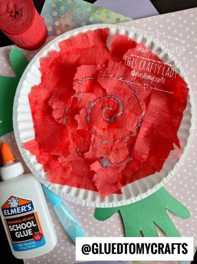 A paper plate decorated with torn red crepe paper in a rose shape, with a green paper hand as a stem. Elmer’s glue, a glue stick, red crepe paper, and blue ribbon are nearby. A pink star-patterned paper is underneath.