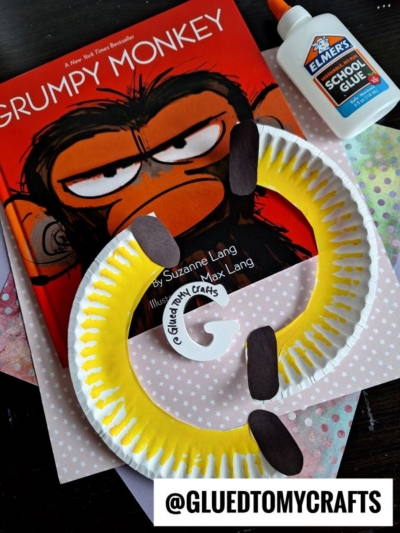 A “Grumpy Monkey” book, Elmer’s School Glue, and a handmade yellow paper plate craft monkey with brown ears and tail on a polka dot background—perfect for summer craft ideas. @GluedToMyCrafts is displayed at the bottom.