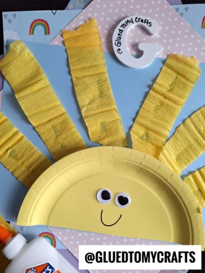 A paper plate painted yellow with googly eyes and a smile, decorated with yellow crepe paper rays to resemble a sun. Craft supplies, rainbows, and a @GLUEDTOMYCRAFTS label are also visible.