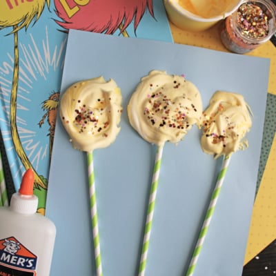 Three DIY truffula trees made from puffy paint on striped paper straws are displayed on blue paper, with craft supplies, sprinkles, glue, and The Lorax book in the background.