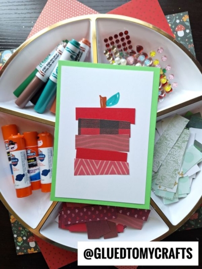 A handmade card featuring a layered paper apple sits on a tray with markers, glue sticks, colorful gems, and patterned paper scraps—perfect inspiration for FALL CRAFTS FOR KIDS. The username @GLUEDTOMYCRAFTS is displayed at the bottom.