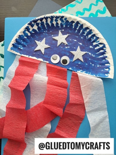 A kids craft jellyfish made from a painted paper plate, decorated with silver stars, two googly eyes, and red and white crepe paper streamers as tentacles, on a blue background. Text reads @GLUEDTOMYCRAFTS.