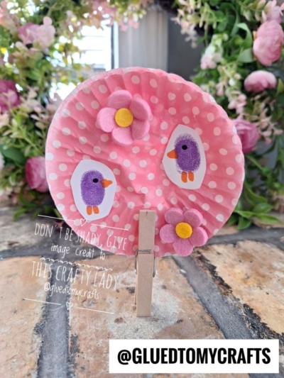 A pink polka dot paper circle decorated with purple bird stickers and flower embellishments is clipped with a clothespin. This charming display, inspired by cherry blossom tree crafts, sits on a brick surface with flowers in the background.