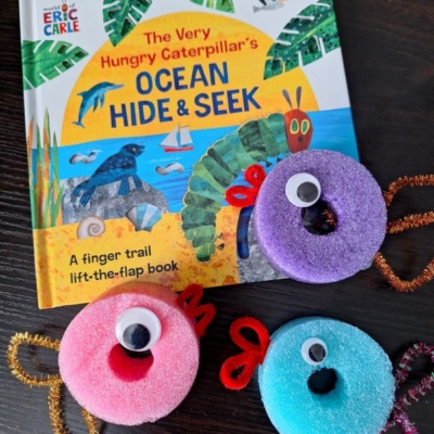 A copy of The Very Hungry Caterpillars Ocean Hide & Seek book is on a table next to three colorful, round, fuzzy crafts with googly eyes and pipe cleaner antennae. @GLUEDTOMYCRAFTS is written at the bottom.