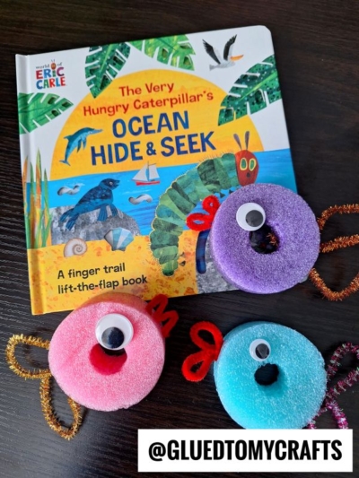 A copy of The Very Hungry Caterpillars Ocean Hide & Seek book is on a table next to three colorful, round, fuzzy crafts with googly eyes and pipe cleaner antennae. @GLUEDTOMYCRAFTS is written at the bottom.