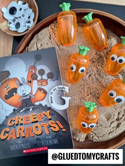 A childrens book titled Creepy Carrots! is placed on a tray with sand and several plastic carrot containers with googly eyes. Paper carrot character crafts and the Instagram handle @GLUEDTOMYCRAFTS are also visible.