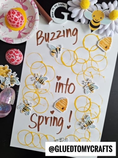 A craft project with the words Buzzing into Spring! surrounded by stamped yellow circles, bee stickers, and honeycomb shapes. Decorative items, including a bee plush, painted egg, and gems, are nearby. @GLUEDTOMYCRAFTS is labeled below.