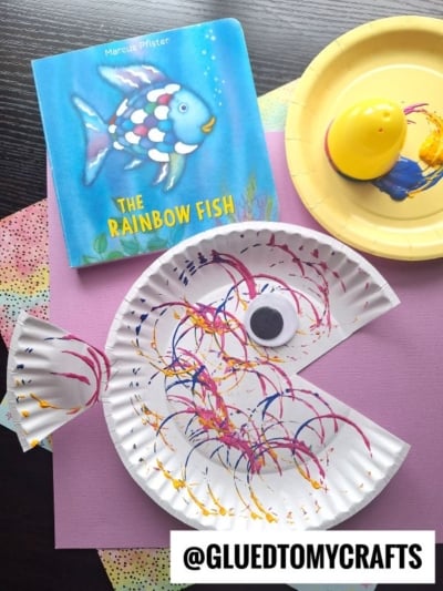 A paper plate decorated like a colorful fish with paint patterns, made using Plastic Egg Stamp Crafts, sits on a pink sheet next to “The Rainbow Fish” book and a yellow paint stamper. The Instagram handle @gluedtomycrafts is visible.