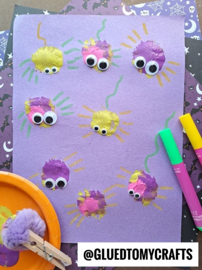 Purple paper with yellow and purple pom-pom painted spiders, each with googly eyes and marker-drawn legs. This fun INSECT CRAFT IDEAS project uses craft supplies, markers, a pom-pom clip, and Halloween-themed paper in the background.