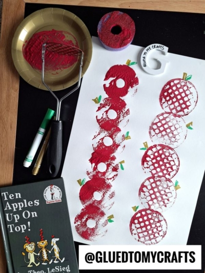 A sheet of paper with red apple prints, a roller with red paint, a gold plate with paint, a green marker, apple-shaped cutouts, and the book Ten Apples Up On Top! by Dr. Seuss on a black surface.