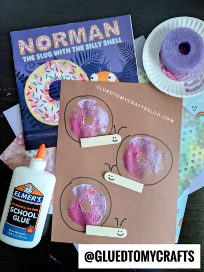 A flat lay of a donut-themed craft project with pink painted donuts on brown paper, a bottle of Elmers school glue, a purple painted sponge, childrens book Norman the Slug with the Silly Shell, and decorative papers.