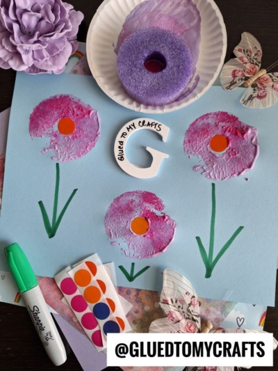 A craft scene with a blue paper showing three pink flower prints with orange centers and green stems, surrounded by a marker, stickers, butterflies, a purple flower, a foam donut, and a “@GLUEDTOMYCRAFTS” label.