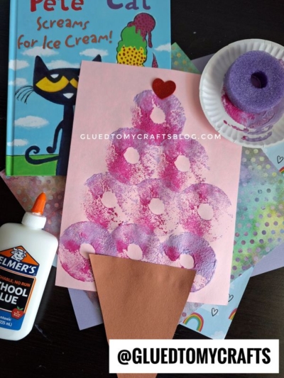 A childrens craft project features a pink ice cream cone made with paint stamps on paper, surrounded by colorful paper, a bottle of Elmers glue, a painted sponge, and a Pete the Cat book.