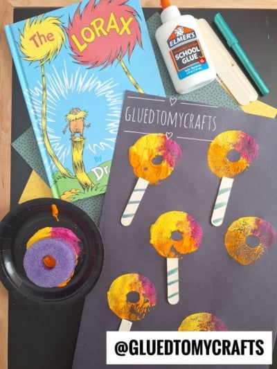 A Dr. Seuss The Lorax book, glue, markers, and a craft project with pool noodle stamped Truffula trees made from paint and popsicle sticks are on a table. A sponge with yellow and purple paint sits by a black paper plate. @GLUEDTOMYCRAFTS is written on the image.