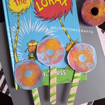 A copy of The Lorax book lies on a table with three handmade Truffula trees crafted from paper and popsicle sticks, and a painted sponge resembling a donut nearby. The handle @GLUEDTOMYCRAFTS is visible on the image.