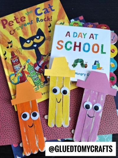 Two children’s books, Pete the Cat: Crayons Rock! and A Day at School, are displayed with three colorful popsicle stick crayon crafts with hats and googly eyes on a patterned surface.