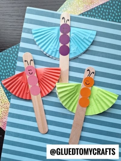 Three butterfly crafts made from wooden popsicle sticks and colored cupcake liners as wings resemble fun cupcake liner card crafts, with paper circles for bodies, drawn smiling faces and antennae, all set against colorful paper backgrounds.