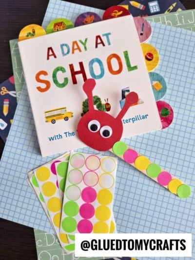 A children’s book titled “A Day at School” with colorful dot stickers and a red Hungry Caterpillar craft made of paper circles and googly eyes, arranged on patterned papers. The text @GLUEDTOMYCRAFTS is at the bottom.