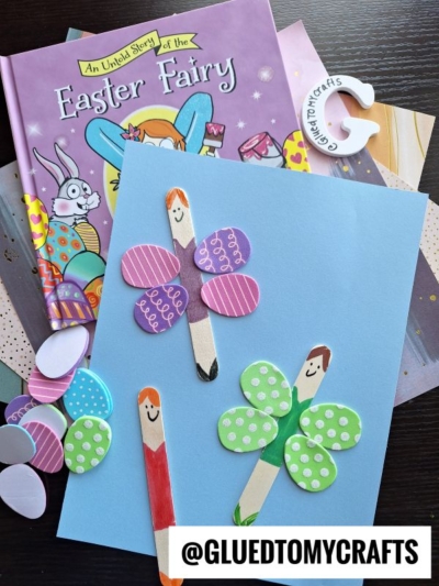 A book titled An Untold Story of the Easter Fairy is next to a blue paper with two craft stick fairies made from decorated paper wings. Extra paper shapes and a @GLUEDTOMYCRAFTS tag are also visible.