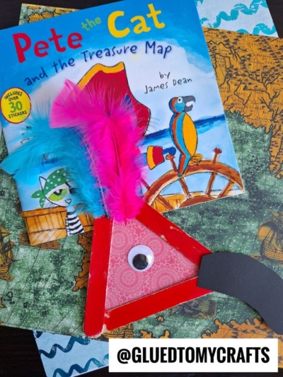 A Pete the Cat “Treasure Map” book with a pirate-themed red paper craft featuring colorful feathers, a googly eye, and a black paper mustache, placed on top of two illustrated map pages. @GLUEDTOMYCRAFTS is at the bottom.