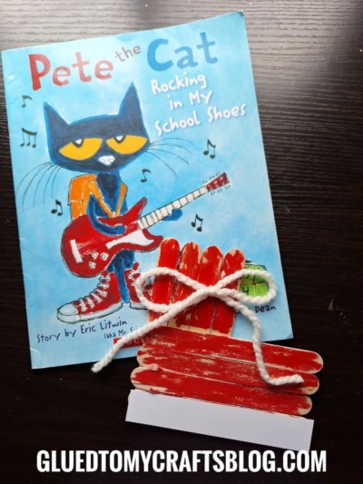 A Pete the Cat: Rocking in My School Shoes book sits on a dark surface next to a red school bus craft made from colored popsicle sticks, white paper, and yarn. The website gluedtomycraftsblog.com is at the bottom.
