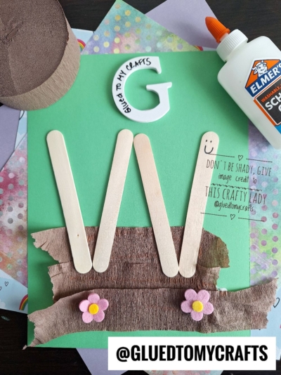 A green paper craft featuring a large letter W made from popsicle sticks, brown paper at the bottom, two pink flower embellishments, a white foam letter G, a bottle of glue, and @GLUETOMYCRAFTS text.