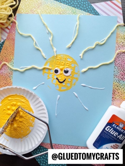 A blue paper with a yellow sun made from paint and yarn. The sun, created as a potato masher stamped craft, has googly eyes, a smiling mouth, and yarn rays. Craft supplies like glue, paint, and a scooper are around the paper.