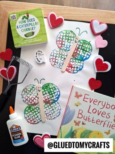 A craft project with two butterfly prints made from paint and popsicle sticks on white paper, surrounded by heart decorations, childrens books about butterflies, glue, and a heart-shaped cookie cutter.