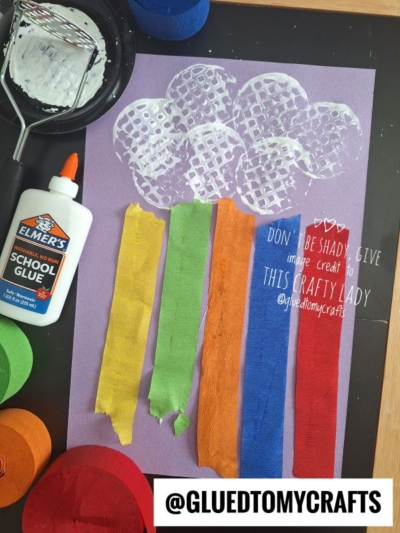 A children’s potato masher stamped craft creates a rainbow with white paint clouds and colorful crepe paper rain on purple paper. A bottle of glue and the paint-covered potato masher sit beside the cheerful project.