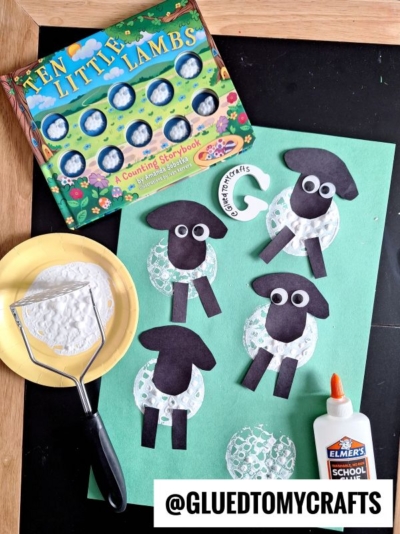 A green paper with lamb crafts made from black and white shapes, googly eyes, and bubble wrap prints. Nearby are glue, a potato masher with white paint, a yellow plate, and the book Ten Little Lambs.