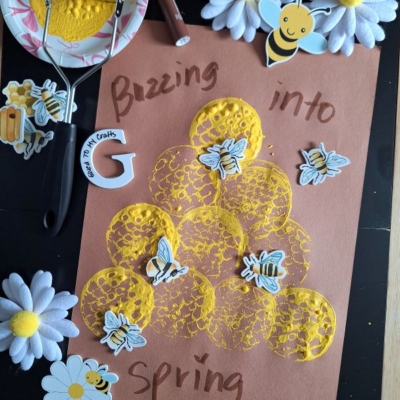 A spring-themed potato masher stamped craft features yellow paint circles, bee stickers, and faux flowers on brown paper with the words Buzzing into spring in marker. The handle @GLUEDTOMYCRAFTS appears at the bottom.