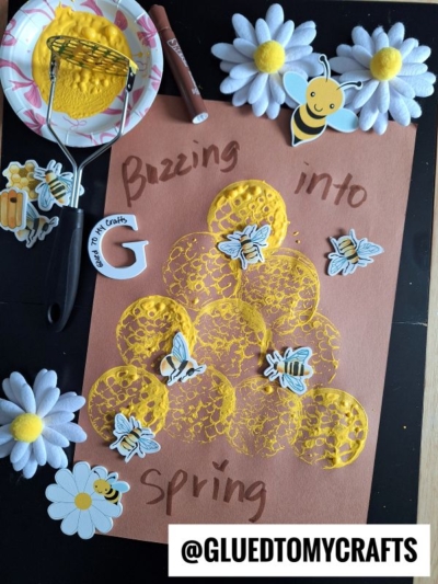 A spring-themed potato masher stamped craft features yellow paint circles, bee stickers, and faux flowers on brown paper with the words Buzzing into spring in marker. The handle @GLUEDTOMYCRAFTS appears at the bottom.