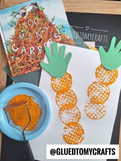 A children’s carrot-themed potato masher stamped craft featuring orange paint-stamped circles for carrot shapes and green paper handprints as tops, displayed beside the book Too Many Carrots and the potato masher used for stamping.