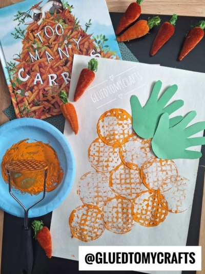 A children’s carrot craft: a book titled “Too Many Carrots,” a potato masher stamped craft with orange circles for carrots, two cut-out green handprints as tops, a paint roller, and faux carrots.
