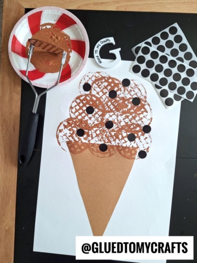 A paper ice cream cone craft with brown paint waffle texture, created using a potato masher stamped craft technique and black dot stickers. Nearby are a spatula with brown paint, a painted cookie, and a sheet of black dot stickers. GLUEDTOMYCRAFTS is labeled at bottom.