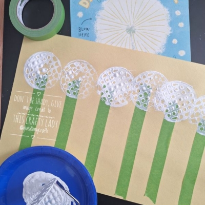 A children’s dandelion craft is shown: stamped white paint dandelions on yellow paper with green stems, a paint-covered jar, a blue plate, and the book “Dandelion Magic.” The handle reads @GLUEDTOMYCRAFTS.
