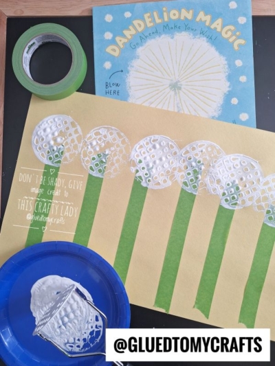 A children’s dandelion craft is shown: stamped white paint dandelions on yellow paper with green stems, a paint-covered jar, a blue plate, and the book “Dandelion Magic.” The handle reads @GLUEDTOMYCRAFTS.