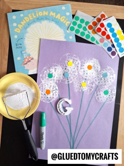 A children’s craft project inspired by the book “Dandelion Magic.” A purple paper shows dandelions made with white paint and dot stickers, surrounded by a paint-covered potato masher, a green marker, and sticker sheets.