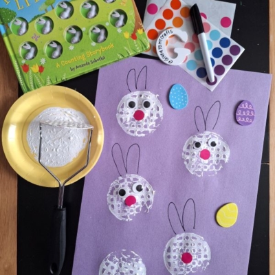 A bunny-themed children’s craft setup: a “Ten Little Bunnies” book, a purple sheet with bunny faces stamped using a potato masher and white paint, colored dot stickers, a black marker, and two painted egg-shaped cutouts.
