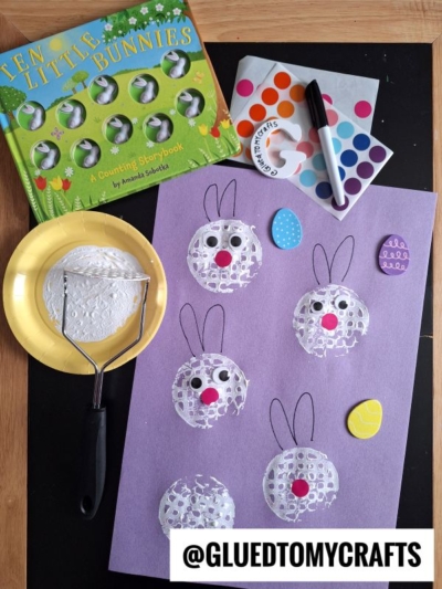 A bunny-themed children’s craft setup: a “Ten Little Bunnies” book, a purple sheet with bunny faces stamped using a potato masher and white paint, colored dot stickers, a black marker, and two painted egg-shaped cutouts.