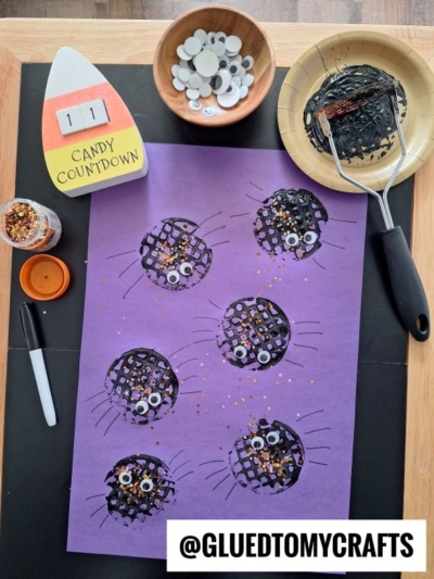 A sheet of purple paper with six black cat faces, created using a potato masher stamped craft, each with googly eyes, whiskers, and gold glitter. Nearby are a glue stick, marker, bowl of googly eyes, paint roller, and a candy countdown sign.